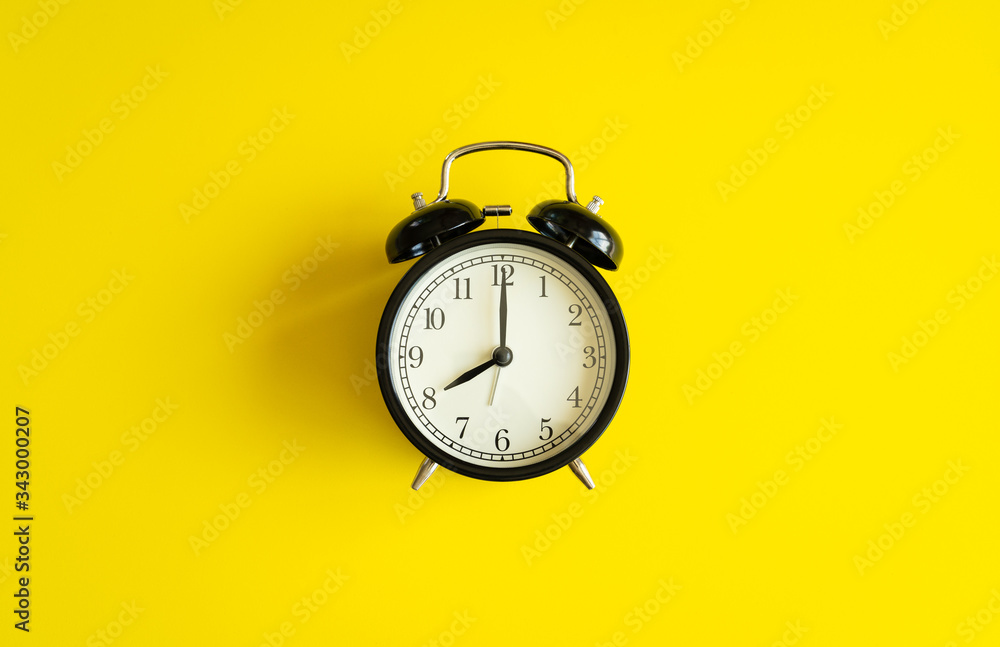 black alarm clock from 7 a.m. to 8 a.m. with bright yellow  background and shadows from the changing light of the sun's movements in morning and working hours concept front shot.