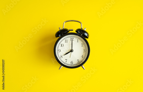 black alarm clock from 7 a.m. to 8 a.m. with bright yellow background and shadows from the changing light of the sun's movements in morning and working hours concept front shot.