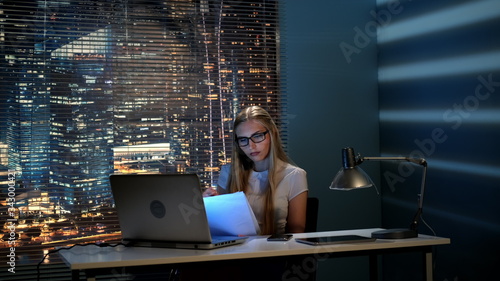 Young female professor correcting her report late at night and checking smartphone for missed calls or new messages