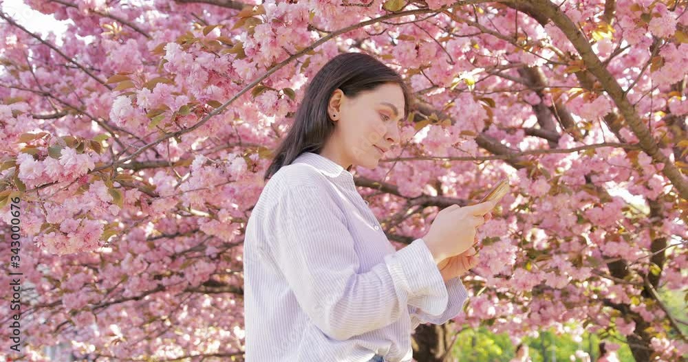 Attractive young woman standing at the sunny park and chatting with friends, using cellphone outdoors, sunset blooming flowers background Attractive young woman standing at the sunny park and chatting with friends, using cellphone outdoors, sunset blooming flowers background