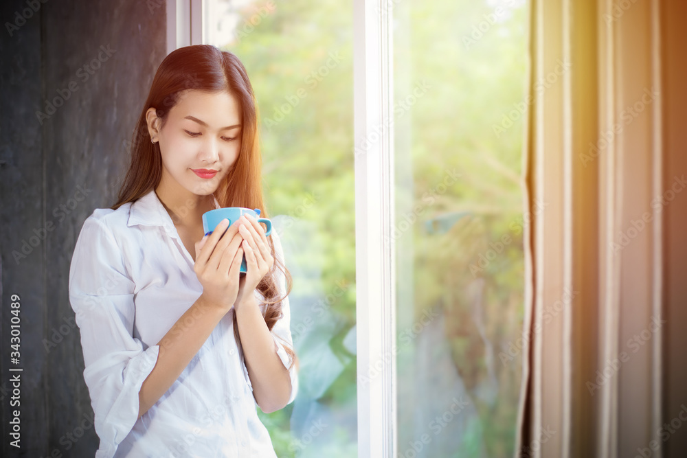 An Asian woman stood holding a coffee cup and opened the curtain on the window after waking up in the morning of the day at home. An Asian woman stood holding a coffee cup and opened the curtain on the window after waking up in the morning of the day at home.