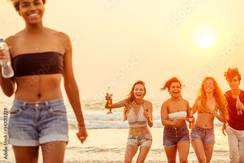 african american woman holding plastic bottle of water , behind her group of mixed race internation friends dancing and having fun with lemonade on sunset beach
