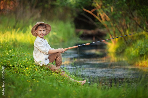 child boy in rustic clothes and a wicker hat sits with a fishing rod in his hands and catches fish on the river bank. hobbies and leisure for men