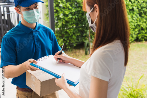 Asian young delivery man courier shopping online with package post box in uniform protective face mask coronavirus he making service woman customer using pen signature on paper book at home, COVID-19