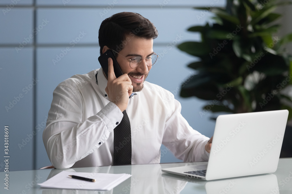 Happy businessman talking on smartphone looking at laptop screen. Smiling successful man manager make online success deal using modern computer. Employee at work. Happy businessman talking on smartphone looking at laptop screen. Smiling successful man manager make online success deal using modern computer. Employee at work.