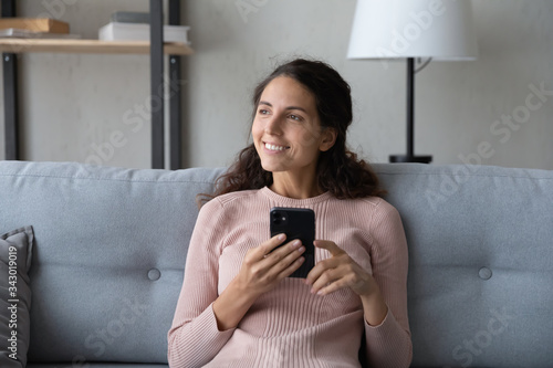 Distracted young smiling woman resting on comfortable sofa with phone in hands, dreaming of summer vacation, visualizing future alone at home. Pleasant happy girl enjoying communicating online.