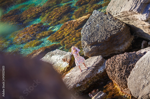 A little pretty girl in a dress sits on a large stone by the sea. A child on a background of turquoise water and rocks, a summer walk on the background of a beautiful landscape