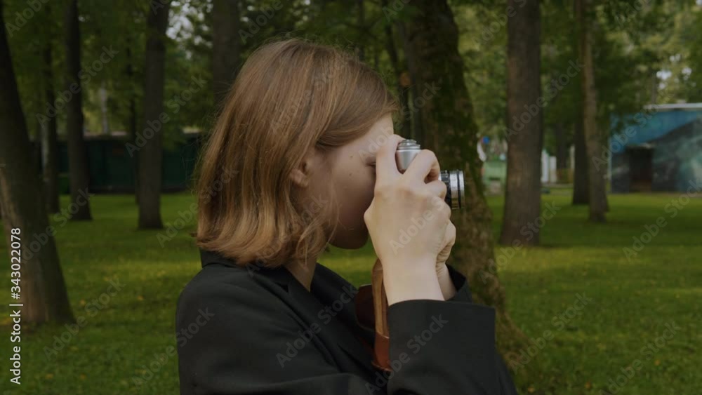 Teenage blonde girl appears in the frame on the left, takes a picture with a vintage film camera and leaves the frame on the right.Against the background of a green park. Teenage blonde girl appears in the frame on the left, takes a picture with a vintage film camera and leaves the frame on the right.Against the background of a green park.