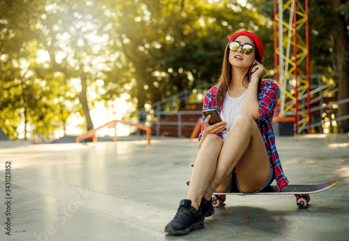 Young stylish woman dressed in youth clothes sits on a skateboard and listens to music on headphones at a skatepark. Summertime