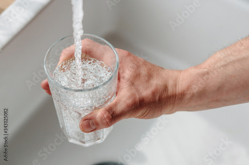 A photograph of a male hand that holds a glass over a sink and pours clean tap water. The photo is lit by sunlight.