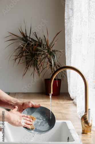 Men's hands wash the plate in the sink. Clean water flows from the tap. The photo is lit by sunlight.