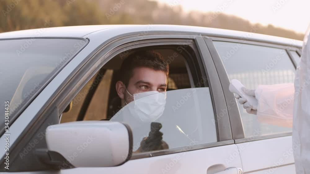 A woman in full-coverage equipment is at the roadblock. She has stopped a car to check the temperature of the passengers if they are not sick. The man driving a car is wearing a disposable mask to A woman in full-coverage equipment is at the roadblock. She has stopped a car to check the temperature of the passengers if they are not sick. The man driving a car is wearing a disposable mask to