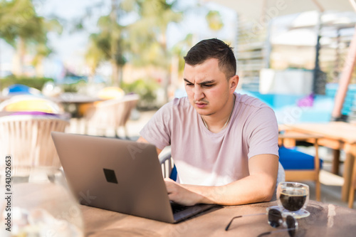 Serious young handsome man with cup of coffee and working with laptop in cafe outdoors
