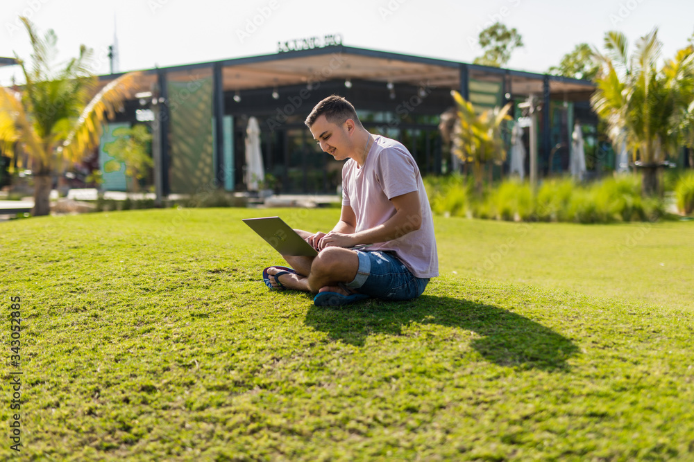 Young smiling man with laptop on grass in city park Young smiling man with laptop on grass in city park