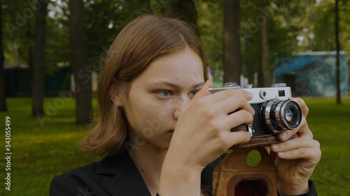 Close-up of a teenage blonde girl who holds a vintage film camera and sets the shutter.Against the background of a green park.