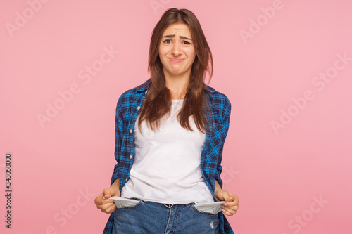 Poor student, unemployment. Portrait of upset jobless girl in checkered shirt turning out empty pockets, showing no money gesture, worried about debts. indoor studio shot isolated on pink background photo