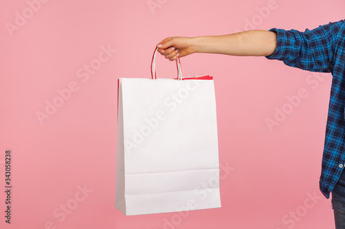 Closeup of female hand in checkered shirt holding shopping bags, showing package with white copy space for store advertising, promotional text or image. indoor studio shot isolated on pink background
