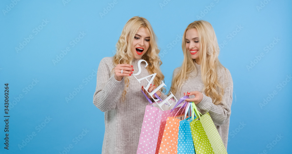 Two bright blondes with color bags in thier hands on a sale, isolated blue neutral background