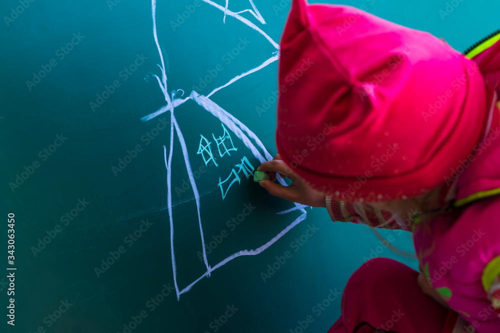 A girl in a pink jacket draws with chalk on a school blackboard a house. In her hands is chalk