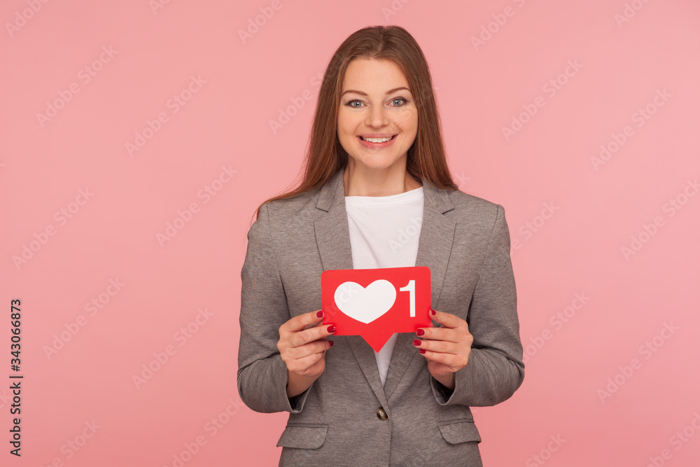 Popularity in social media. Portrait of elegant woman in business suit holding network heart Like icon and smiling to camera, recommending to follow blog. studio shot isolated on pink background Popularity in social media. Portrait of elegant woman in business suit holding network heart Like icon and smiling to camera, recommending to follow blog. studio shot isolated on pink background