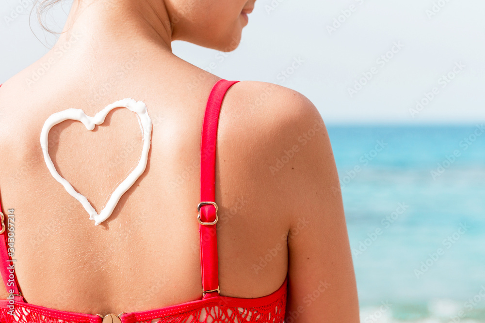 Fototapeta premium Beautiful girl in red swimsuit has sunblock in shape of heart at the back at the beach