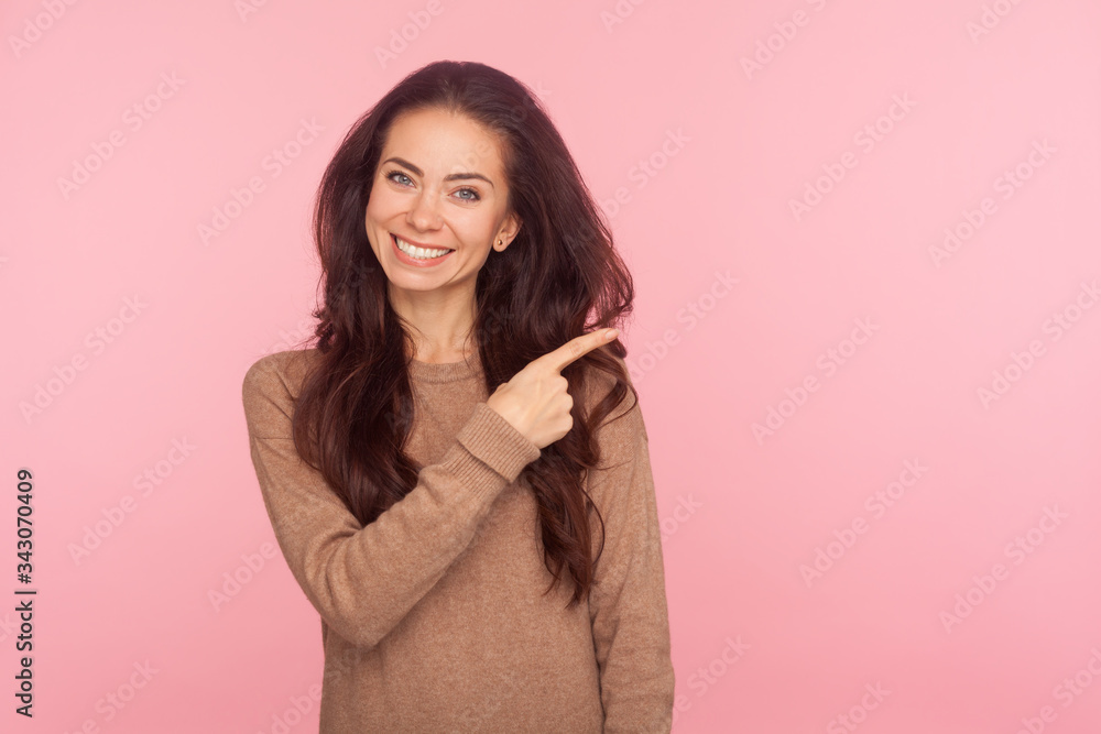 Attention to advertising! Portrait of cheerful young woman with brunette wavy hair pointing aside, showing copy space for commercial text, blank wall with idea presentation. studio shot isolated Attention to advertising! Portrait of cheerful young woman with brunette wavy hair pointing aside, showing copy space for commercial text, blank wall with idea presentation. studio shot isolated