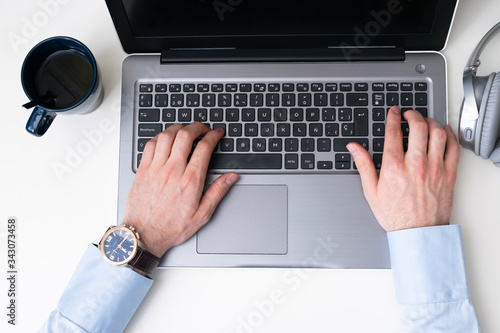 Man typing a laptop keyboard while working, he has tea cup on top left corner and headphone on top right corner. Top point of view
