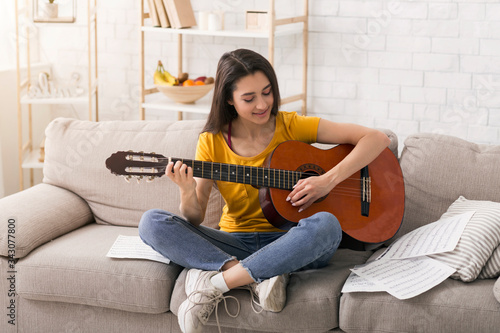 Stay home, have fun. Lovely musical girl with notes playing acoustic guitar on comfy sofa indoors