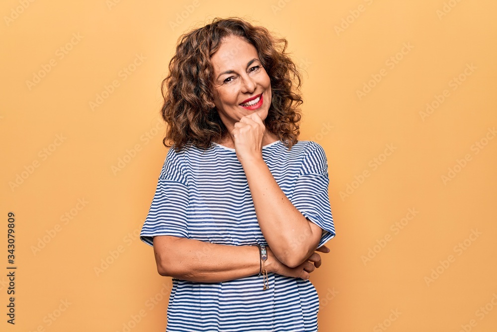 Middle age beautiful woman wearing striped t-shirt standing over isolated yellow background smiling looking confident at the camera with crossed arms and hand on chin. Thinking positive. - obrazy, fototapety, plakaty 