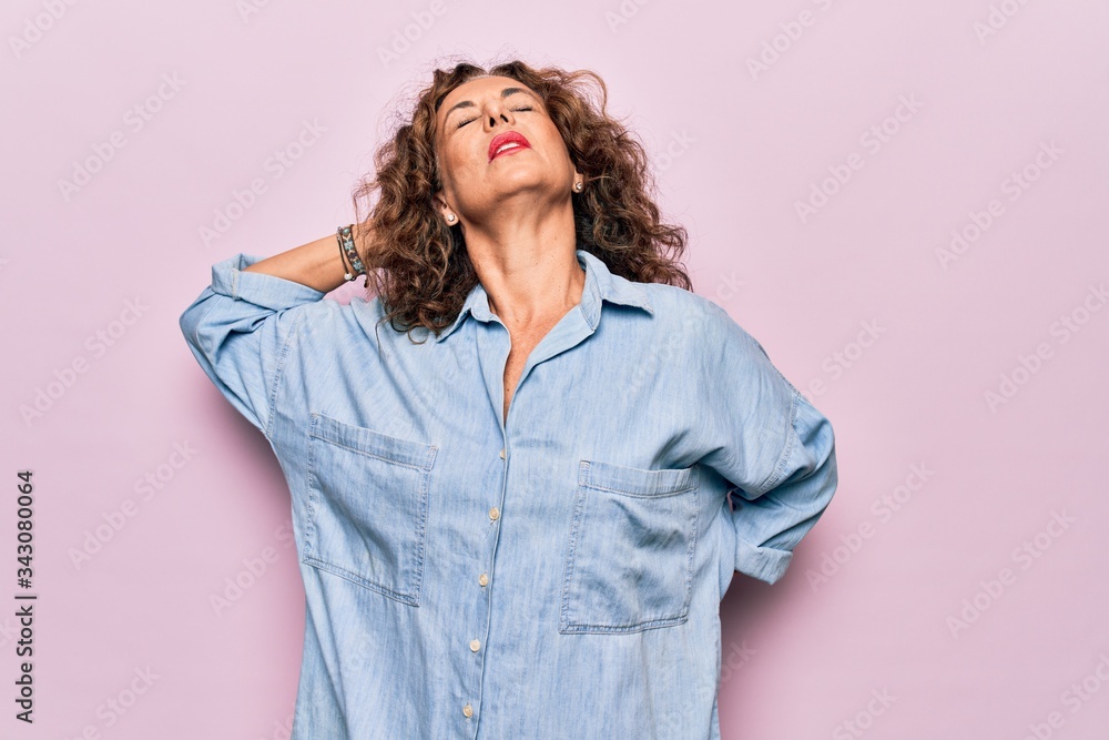Middle age beautiful woman wearing casual denim shirt standing over pink background Suffering of neck ache injury, touching neck with hand, muscular pain Middle age beautiful woman wearing casual denim shirt standing over pink background Suffering of neck ache injury, touching neck with hand, muscular pain