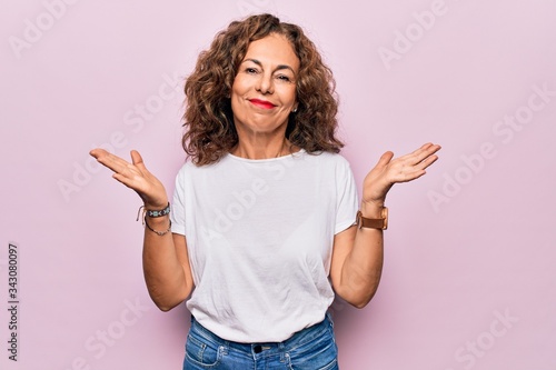 Middle age beautiful woman wearing casual t-shirt standing over isolated pink background clueless and confused with open arms, no idea and doubtful face. photo