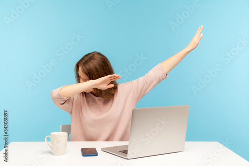 Dabbing trends. Overjoyed woman showing dab dance gesture, performing internet meme of success, sitting at workplace with laptop, home office job. indoor studio shot isolated on blue background photo