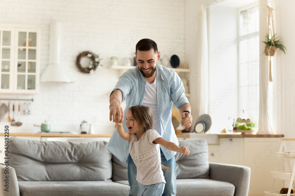 Fototapeta premium Active young father holding hands of cute little preschool kid daughter, dancing to energetic pop music in modern living room. Happy different generations family having fun together at home.