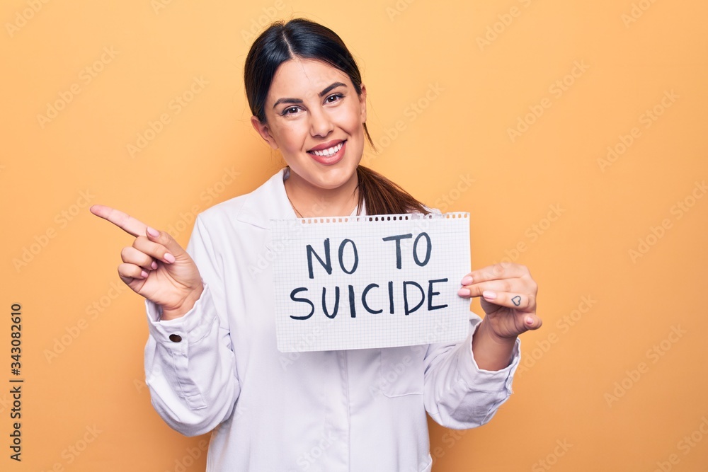 Young beautiful brunette psychologist woman holding paper with not to suicide message smiling happy pointing with hand and finger to the side