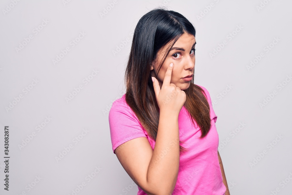 Young beautiful brunette woman wearing casual pink t-shirt standing over white background Pointing to the eye watching you gesture, suspicious expression Young beautiful brunette woman wearing casual pink t-shirt standing over white background Pointing to the eye watching you gesture, suspicious expression