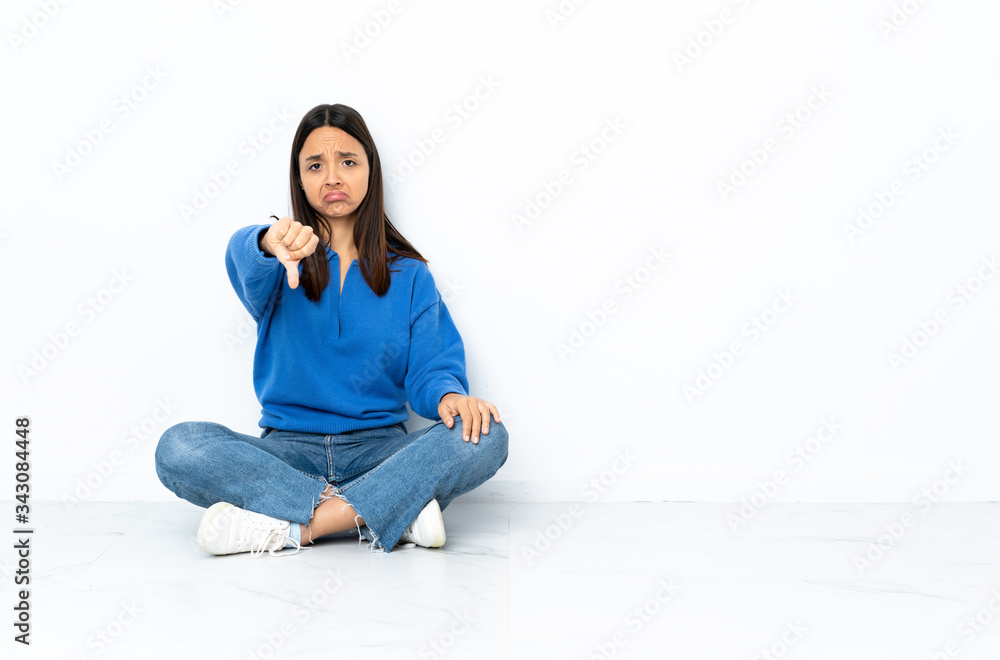 Young mixed race woman sitting on the floor isolated on white background showing thumb down with negative expression Young mixed race woman sitting on the floor isolated on white background showing thumb down with negative expression