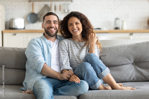 Smiling young bearded man sitting on sofa, cuddling attractive joyful wife, looking at camera. Happy family couple watching comedian movie funny video, enjoying holiday weekend time together at home.