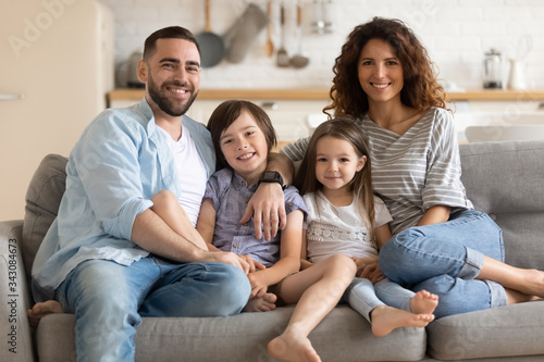 Portrait of happy married couple embracing little kids son daughter, sitting together on comfortable couch in modern studio living room. Smiling cheerful spouses posing for photo with children. © fizkes