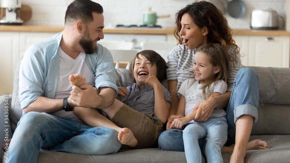 Happy young father tickling little kid boy son while smiling mother cuddling small daughter on couch. Cheerful full family of four having fun on sofa, enjoying weekend holiday time together at home.