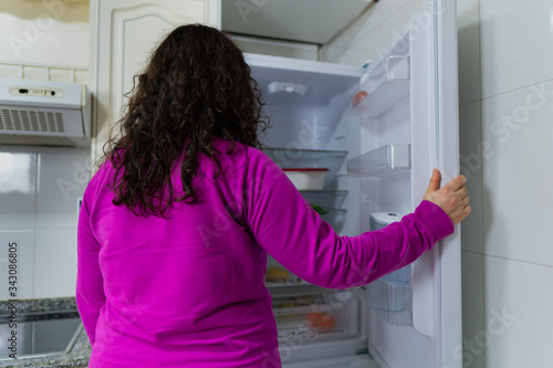 Señora blanca de edad media abre la nevera vacía sin comida debido a la crisis económica. La nevera es blanca y tiene un jersey  violeta.