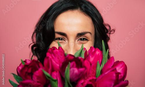 The 8th of March. Close-up photo of a cheerful brunette woman, who is looking in the camera and hiding a part of her face over the bunch of tulips. #343086832