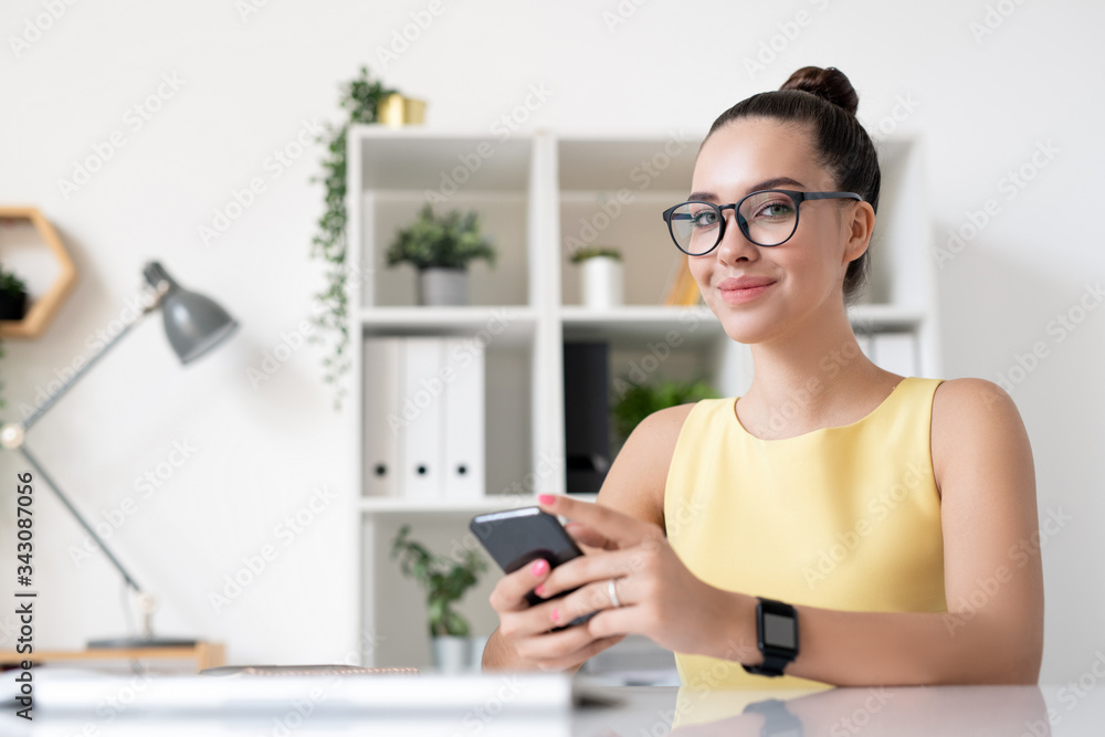 Portrait of content young marketing specialist in yellow dress sitting with smartphone in modern office Portrait of content young marketing specialist in yellow dress sitting with smartphone in modern office