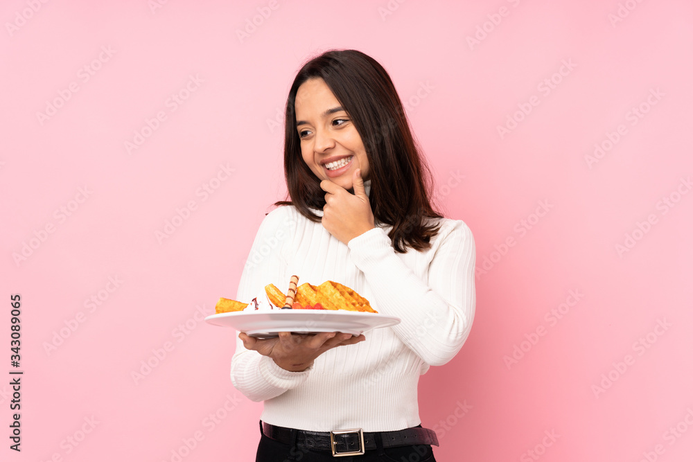Young brunette woman holding waffles over isolated pink background looking to the side Young brunette woman holding waffles over isolated pink background looking to the side