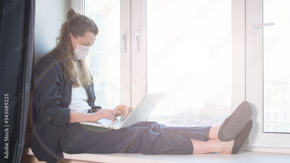Woman wearing mask and working on laptop from home due to coronavirus self isolation. Sitting on window sill. Authentic home workplace. Coronavirus outbreak 2020. Woman wearing mask and working on laptop from home due to coronavirus self isolation. Sitting on window sill. Authentic home workplace. Coronavirus outbreak 2020.