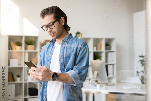 Serious busy young brunette man in glasses checking phone message in own office