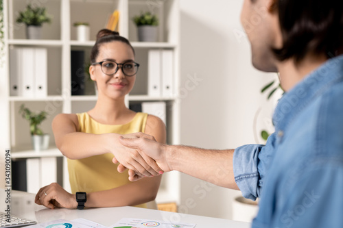 Over shoulder view of man making handshake with attractive businesswoman after meeting in office, startup partnership concept