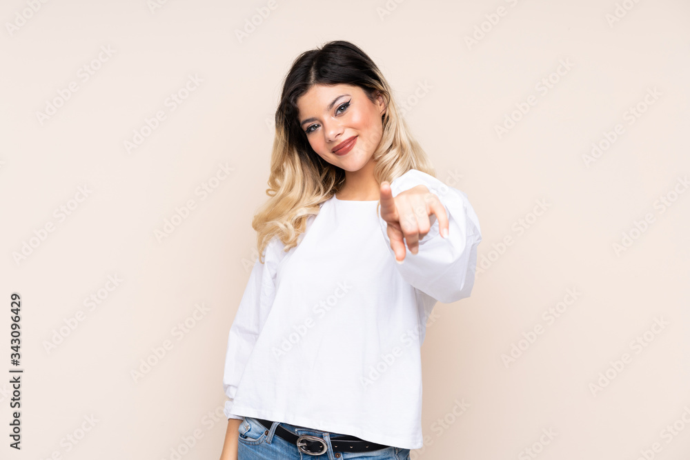 Teenager girl isolated on beige background points finger at you with a confident expression Teenager girl isolated on beige background points finger at you with a confident expression