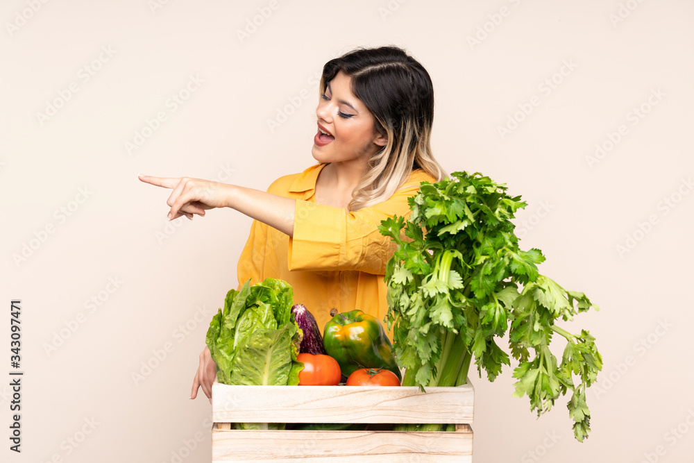 Teenager farmer girl with freshly picked vegetables in a box isolated on beige background pointing finger to the side Teenager farmer girl with freshly picked vegetables in a box isolated on beige background pointing finger to the side