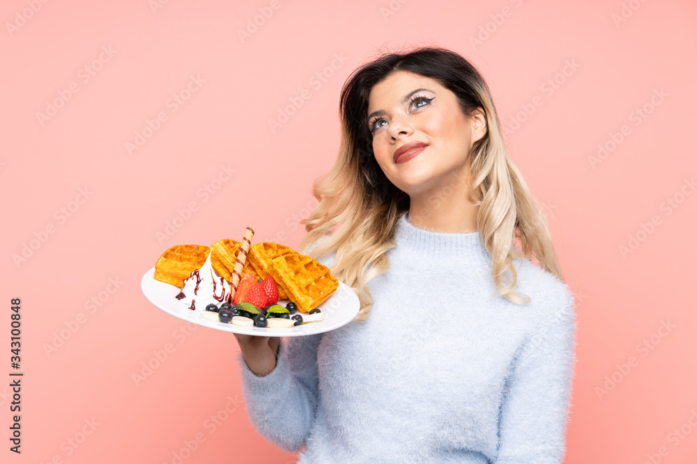 Teenager girl holding waffles on isolated pink background looking up while smiling Teenager girl holding waffles on isolated pink background looking up while smiling