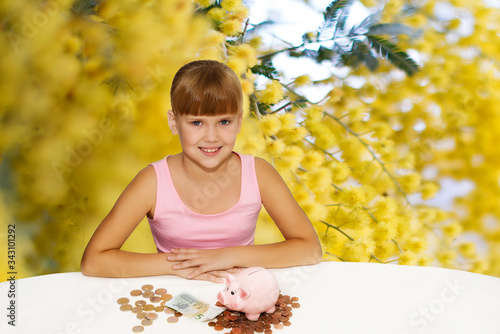 Little lovely girl with coinsand piggy bank on the desk over yellow flowers background. Saving Money for Education Fund. People  children  business concept